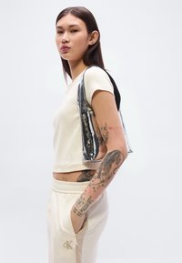 Κρεμ τοπ με κοντά μανίκια και κοντό σχεδιασμό, σετ με κρεμ παντελόνι με λογότυπο CK. Διαφανής τσάντα ώμου με μαύρες λεπτομέρειες.