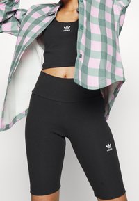 Kvinna som bär svart Adidas crop top och cykelbyxor med en rosa och grön rutig skjorta över, poserande mot en vit bakgrund.