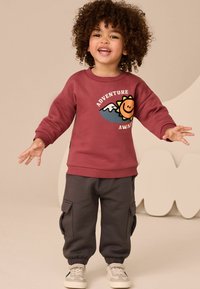 Bordeauxroter Sweatshirt mit einem Sonnen-Graphic und Text, kombiniert mit dunkelgrauen Cargo-Hosen mit Seitentaschen und elastischen Bündchen.