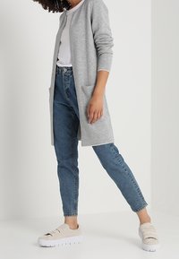 Ljusgrå lång cardigan i mjukt material med långa ärmar och framfickor, parat med åtsittande mörkblå jeans och beige plattformssneakers.
