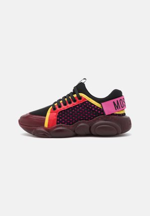 Sneaker nera con accenti rossi, gialli e rosa, lati in mesh strutturato, suola spessa e cinturino rosa con testo nero sul tallone.