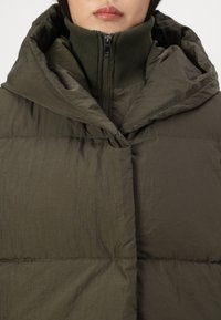 Bunda s kapucňou v olivovej zelenej farbe, s vysokým golierom a zipsom, prešívaná textúra a ľahký materiál. Hladký, matný povrch s jemným leskom.