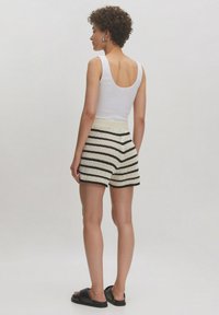 Witte aansluitende tanktop, crèmekleurige haakshorts met zwarte strepen, elastische tailleband, getextureerd patroon, platte zwarte sandalen, model vanuit de achterkant bekeken.