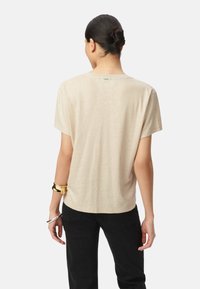 T-shirt beige a maniche corte con una texture morbida e leggera; colletto girocollo semplice; indossato con pantaloni neri; braccialetti metallici visibili sui polsi.