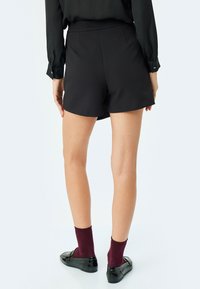 Zwarte shorts met een getailleerd ontwerp, featuring een middelhoge taille, een gladde textuur en twee zijzakken. G gedragen met zwarte instappers en bordeauxrode sokken.
