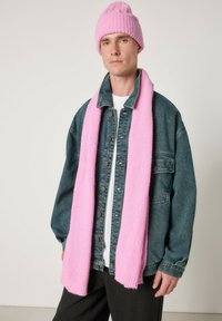 Rausvos spalvos megzta kepurė ir šalikėlis derinami su mėlyna džinsine striuke ant baltos palaidinės. Šalikėlis yra siauras ir ilgas, suteikiantis tekstūros ir šilumos.