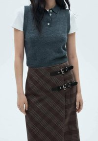 Polo sans manches en maille grise sur une chemise blanche à manches courtes, assorti avec une jupe à carreaux bruns ornée de boucles noires.