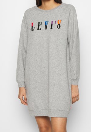 Kobieta w szarej sukience dresowej z długim rękawem i wielokolorowym logo "LEVI'S" na klatce piersiowej, stojąca na jednolitym tle.