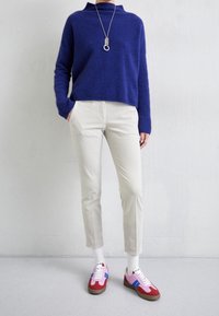 Königsblauer gestrickter Pullover, cremefarbene, schmal zulaufende Hose und mehrfarbige Sneaker (rot, rosa, blau) mit weißen Schnürsenkeln und Akzenten. Silberne Anhänger-Halskette.