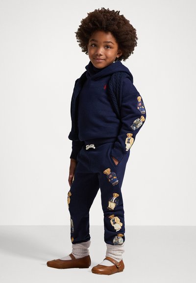 Σκούρο μπλε φούτερ και φόρμες με εκτυπώσεις αρκούδων στα μανίκια και τα πόδια. Καφέ παπούτσια με γκρι κάλτσες. Λείο υλικό και άνετη εφαρμογή.