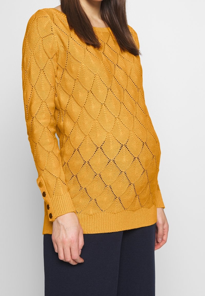 Femme portant un pull en tricot jaune moutarde avec un motif en diamant et des détails de boutons sur le poignet de la manche, associée à un pantalon foncé.