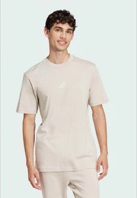 Ανοιχτό μπεζ βαμβακερό T-shirt με κοντά μανίκια και στρογγυλή λαιμόκοψη. Διαθέτει ένα μικρό, ανακλαστικό λογότυπο μάρκας στο στήθος. Απλός σχεδιασμός.