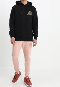 Person som bär en svart hoodie med guldlogo, ljusrosa joggers med cirkulär logo och röda sneakers som står mot en vit bakgrund.