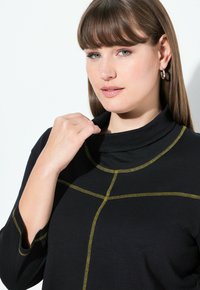 Vrouw met lichte huid en lang bruin haar, gekleed in een zwarte top met gele steken, die de kraag met haar rechterhand vasthoudt, neutrale uitdrukking.