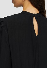 Schwarzes, strukturiertes Kleid mit Knopfverschluss hinten und Schlitzöffnung. Lockere Passform mit subtilen Faltendetails. Weiches, gerafftes Material.