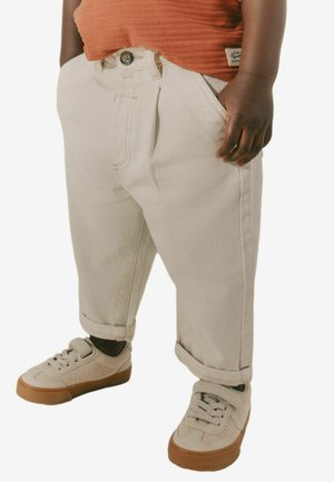 Beige, gefaltete Denim-Hosen mit umgeschlagenen Säumen und Knopfverschluss. Kombiniert mit hellgrauen Sneakers mit strukturiertem Obermaterial und Gummisohle.