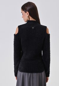 Maglione nero a coste realizzato in materiale morbido con spalle scoperte. Presenta una vita aderente e un piccolo logo sulla schiena. Maniche lunghe.