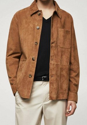 Man in een bruine suède knopenjack over een zwart V-hals T-shirt, gecombineerd met off-white broeken en een zwarte riem.