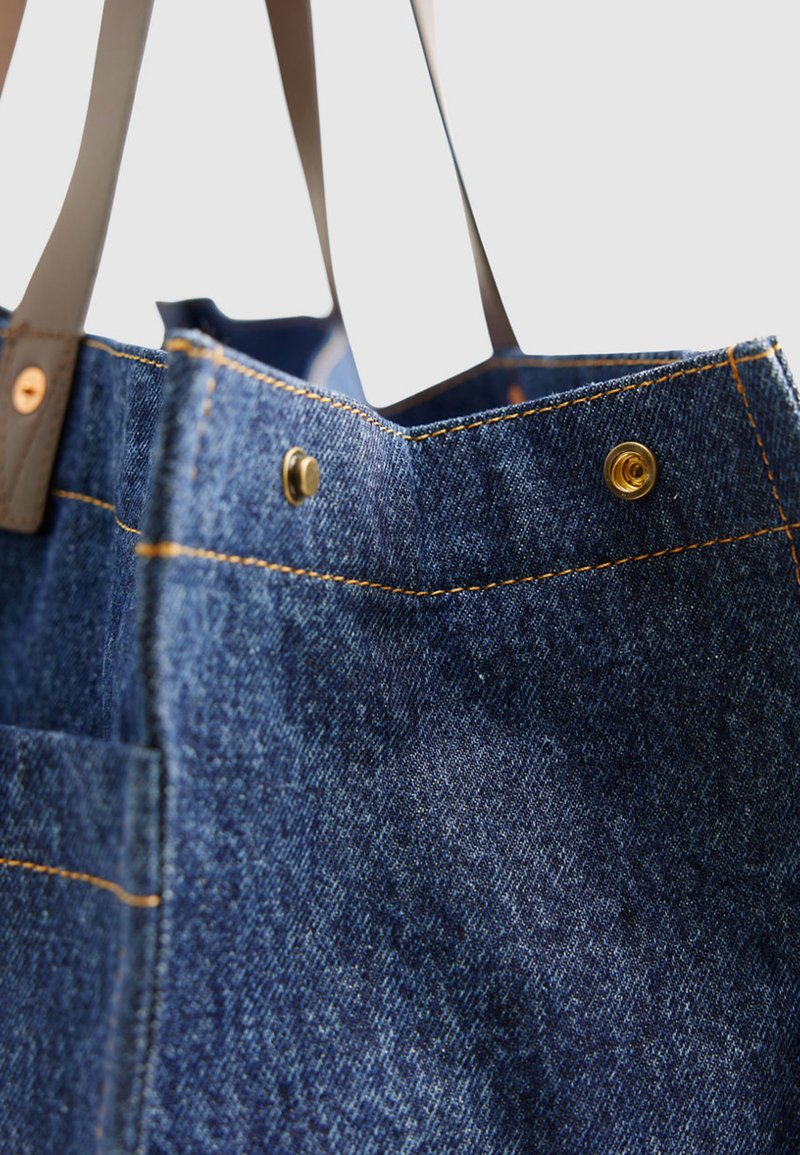 Jeanstasche mit dunkelbraunen Ledergriffen, sichtbaren Messing-Druckknöpfen und gelber Kontrastnaht auf strukturierter Stoffoberfläche.