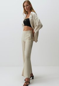 Blazer en lin crème sur un crop top noir, associé à un pantalon évasé crème avec des coutures verticales ; le mannequin porte des sandales à talons noires.