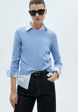 Light blue knit sweater layered over a white collared shirt, paired with dark jeans. Features ribbed cuffs and a casual fit. Dark sunglasses worn.