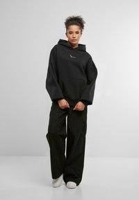 Zwart oversized hoodie met een kangoeroezak en geborduurd logo op de borst, gecombineerd met baggy zwarte cargobroeken en witte sneakers.