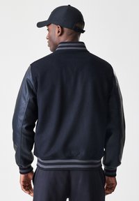 Homem a usar uma jaqueta varsity preta com mangas de couro e um boné preto correspondente, visto de costas contra um fundo claro e liso.