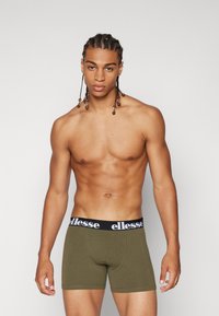 Olivgrüne Boxershorts mit einem schwarzen elastischen Bund, der mit "ellesse"-Branding versehen ist. Der Stoff ist glatt, das Design enganliegend und hat eine Länge bis zur Mitte des Oberschenkels.