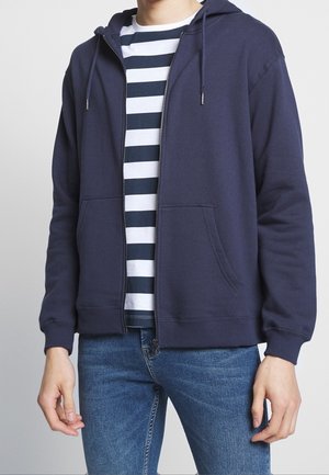 Person iført en navyblå lynlåshoodie over en hvid og navystribet skjorte med blå jeans, stående mod en ensfarvet baggrund.