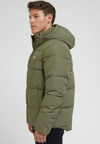 Oliwkowa kurtka puchowa z kapturem, z pikowanym wzorem, z zapięciem na zamek błyskawiczny i przednimi kieszeniami. Wykonana z lekkiego, wodoodpornego materiału.