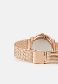 Roségoldenes Mesh-Armbanduhr mit einer polierten Metallverschluss, ausgestattet mit einem minimalistischen runden Zifferblatt und einem glatten, reflektierenden Gehäuseboden.