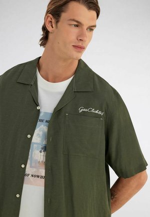 Joven con una camisa de manga corta verde oscuro abierta sobre una camiseta blanca con gráfico, mirando ligeramente hacia su derecha contra un fondo liso.