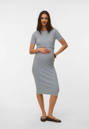 Mujer con un vestido ajustado hasta la rodilla, de manga corta y rayas negras y blancas, con una mano en el vientre, de pie y sonriendo.