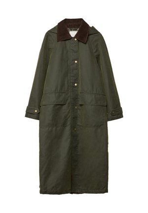 Personne portant un long manteau vert foncé, un pull noir, une jupe blanche ornée de dentelle et des bottes noires montantes sur un fond gris uni.