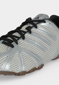 Ασημί αθλητικό παπούτσι Adidas με μαύρα κορδόνια, διχτυωτό πάνω μέρος και καφέ λαστιχένια σόλα σε κοντινή γωνία.