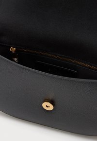 Musta teksturoitu nahkalaukku, jossa on avonainen läppä, joka näyttää mustan kankaan sisustuksen ja kultaisen vetoketjupussin sisäpuolella.