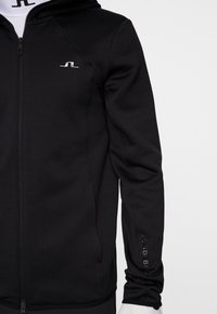 Svart zip-jacka i mjukt material, med huva, logotypaccent i silver och textdetalj längs ärmen.