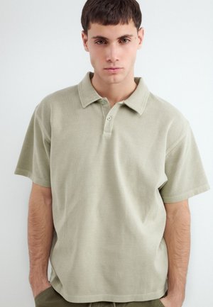 Young man with short dark hair wearing a light beige short-sleeve polo shirt standing against a plain light background.