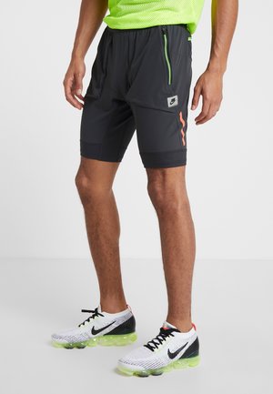 Homme portant un short Nike noir avec une fermeture éclair verte et une bande orange, des chaussures de course Nike blanches avec des semelles translucides vertes.