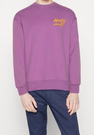 Personne portant un sweat-shirt violet à manches longues avec un petit motif jaune sur la poitrine et un pantalon bleu foncé.
