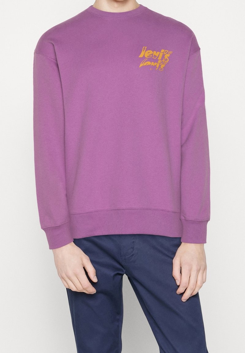 Personne portant un sweat-shirt violet à manches longues avec un petit motif jaune sur la poitrine et un pantalon bleu foncé.