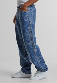 Blue denim jeans with a faded pattern, loose fit, and visible seams, paired with white athletic sneakers.