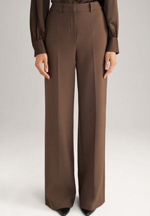 Pantalon marron droit avec plis à l’avant, porté avec un chemisier marron assorti et des chaussures noires pointues sur un fond neutre.