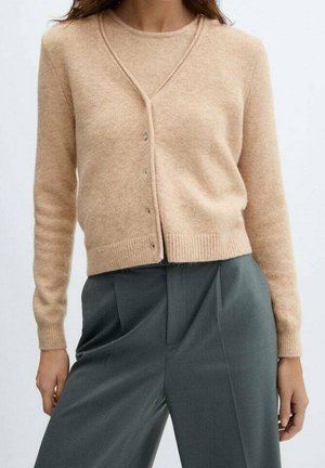 Femme portant un cardigan beige boutonné et un pantalon plissé gris foncé taille haute, les mains détendues le long du corps.