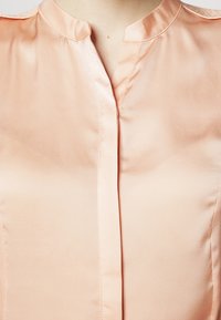 Persikan satiin blouse dengan kerah klasik, tekstur halus, dan penutup depan tersembunyi untuk tampilan yang bersih.