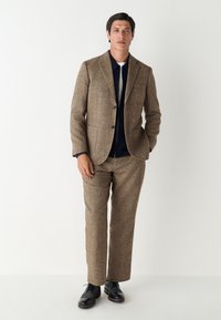 Costume marron texturé à la coupe ajustée, comprenant une veste à revers cranté, deux poches avant, et des chaussures en cuir noir. Chemise bleue marine visible en dessous.