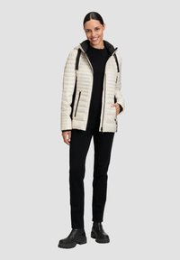 Giacca trapuntata beige chiaro con dettagli neri, cappuccio, zip frontale e tasche laterali, abbinata a un top e pantaloni neri e stivali neri chunky.
