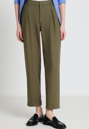 Pantaloni plissettati verde oliva a vita alta con orli risvoltati indossati con una camicia blu abbottonata e mocassini neri lucidi.