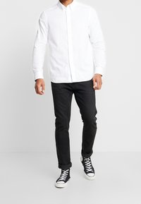 Camisa blanca de botones, de manga larga, combinada con jeans oscuros y zapatillas negras de caña alta. Diseño sencillo, la tela parece suave y nítida.