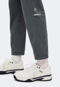 Λευκά sneakers Fila με μαύρες σόλες, φορεμένα με γκρι παντελόνια και άσπρες κάλτσες πάνω από τον αστράγαλο, σε απλό φωτεινό φόντο.
